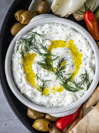 Bowl of creamy yogurt and feta dip, garnished with dill, with chopped vegetables and pita on the side.
