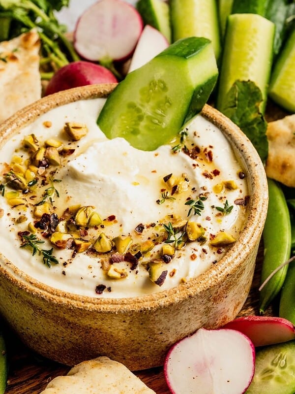 Whipped feta with honey and pistachios in a bowl, garnished with thyme and olive oil, surrounded by pita and sliced vegetables.