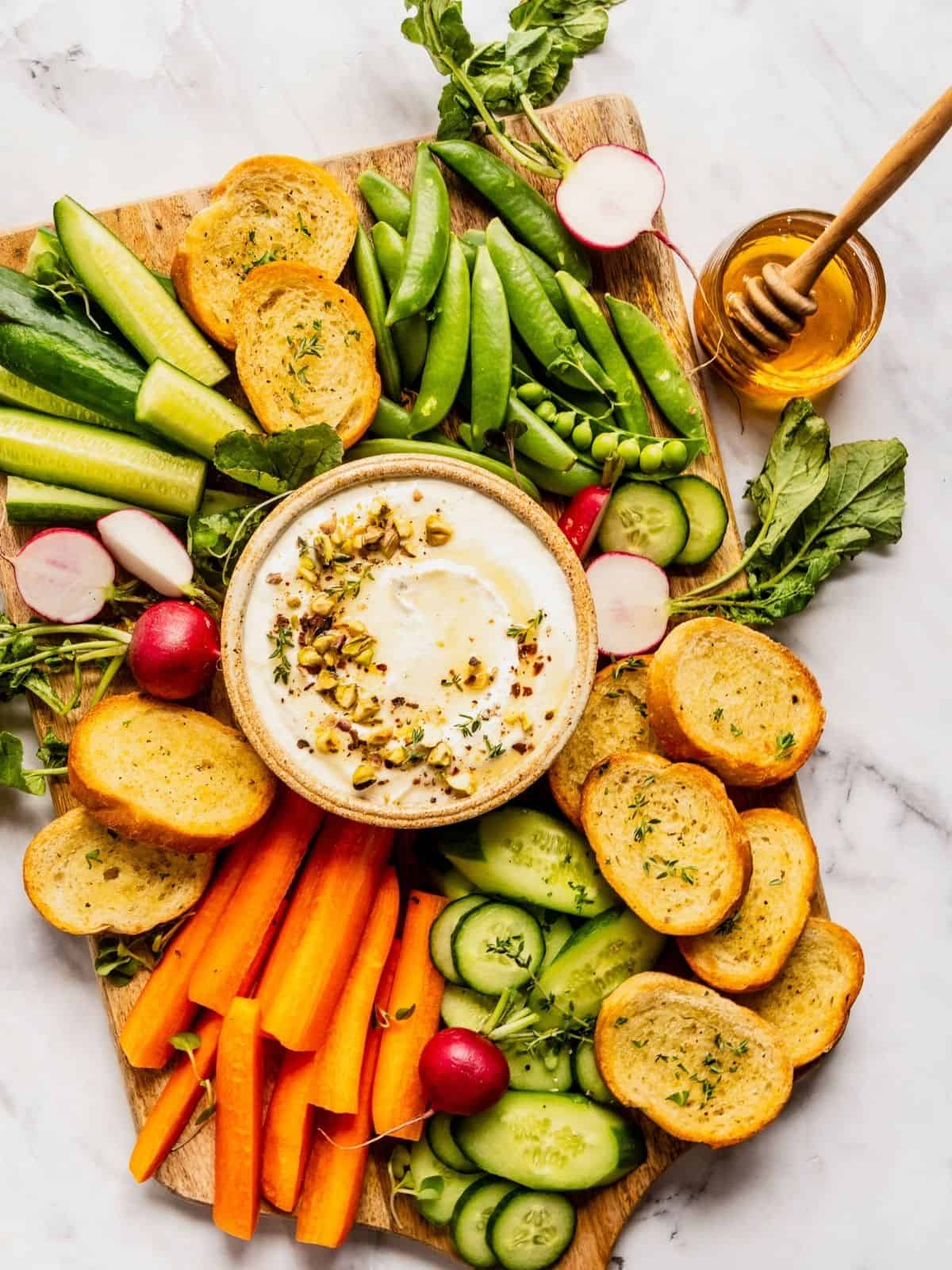 Whipped feta with honey and pistachios in a bowl, garnished with thyme and olive oil, surrounded by pita and sliced vegetables.