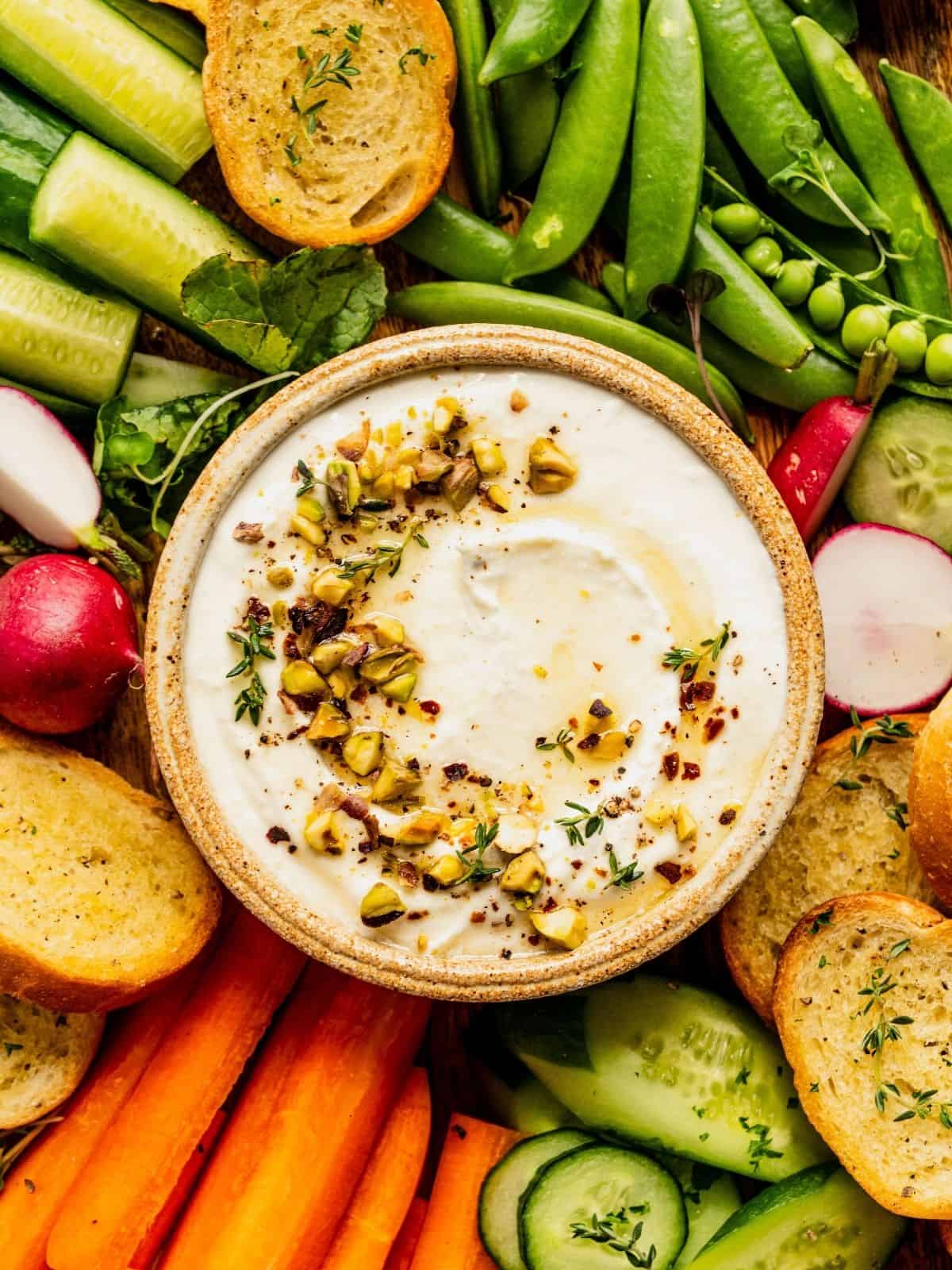 Whipped feta with honey and pistachios in a bowl, garnished with thyme and olive oil, surrounded by pita and sliced vegetables.
