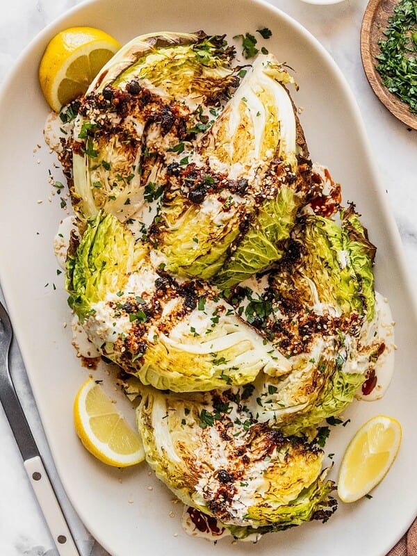 Roasted cabbage wedges with tahini and chili crisp drizzle on a serving platter garnished with parsley and lemon slices.