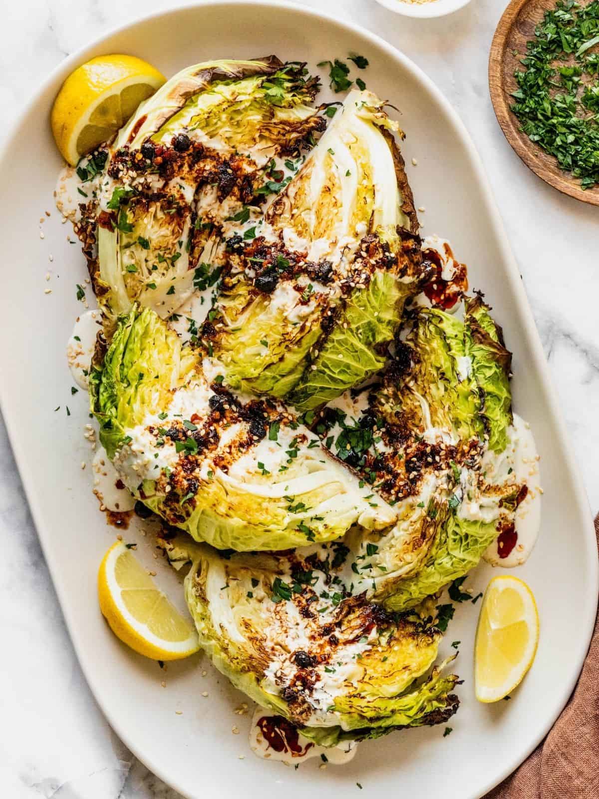 Roasted cabbage wedges with tahini and chili crisp drizzle on a serving platter garnished with parsley and lemon slices.