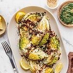 Roasted cabbage wedges with tahini and chili crisp drizzle on a serving platter garnished with sesame seeds, parsley and lemon slices.