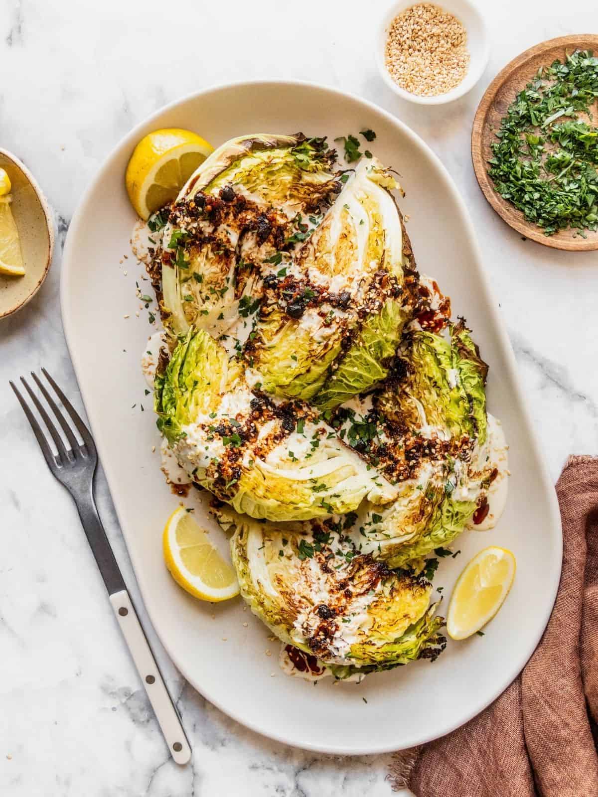 Roasted cabbage wedges with tahini and chili crisp drizzle on a serving platter garnished with sesame seeds, parsley and lemon slices.