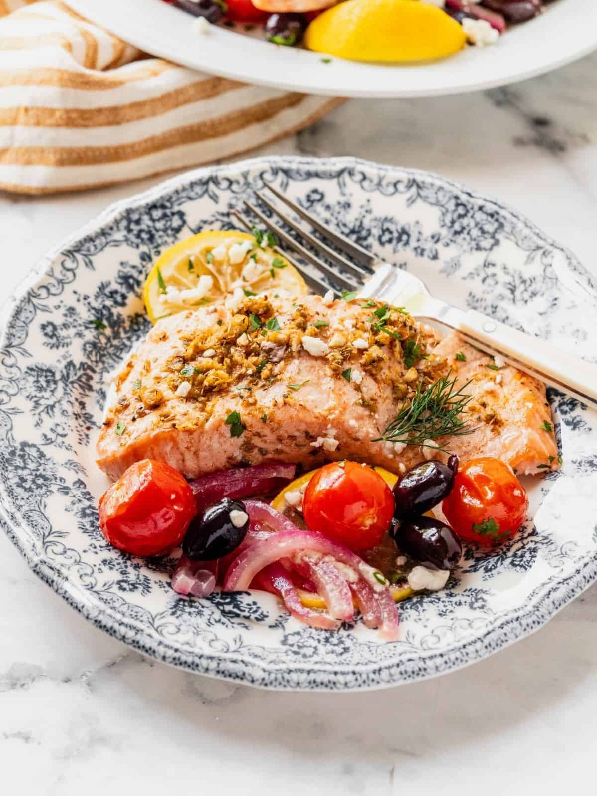 Greek salmon filets served on plates with a side of cherry tomatoes, olives, onion, and lemon, garnished with feta.