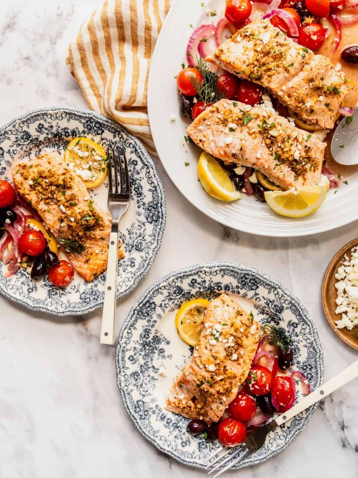 Greek salmon filets served on plates with a side of cherry tomatoes, olives, onion, and lemon.