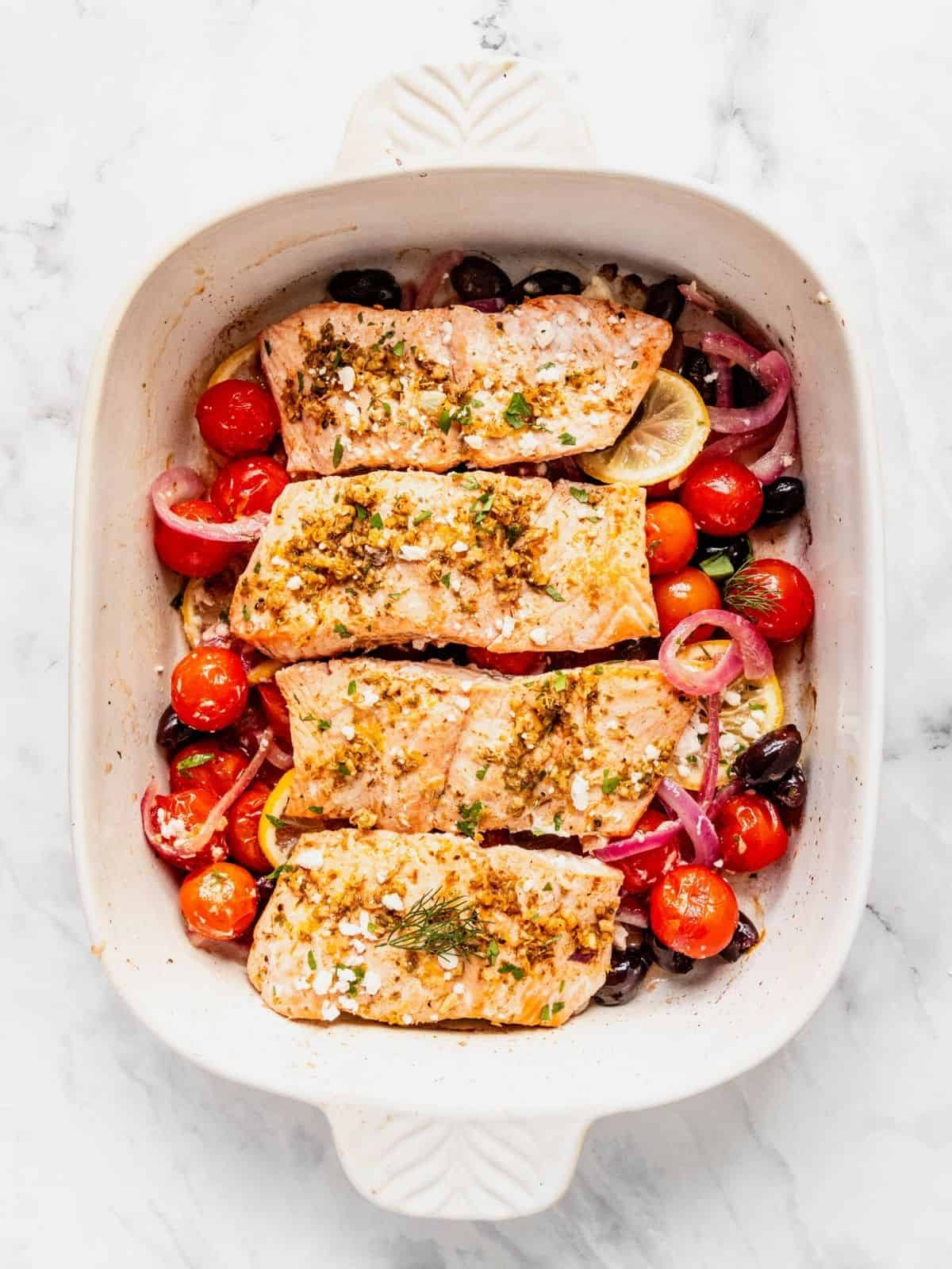 Baked fish fillets with cherry tomatoes, olives, and herbs in a white baking dish.