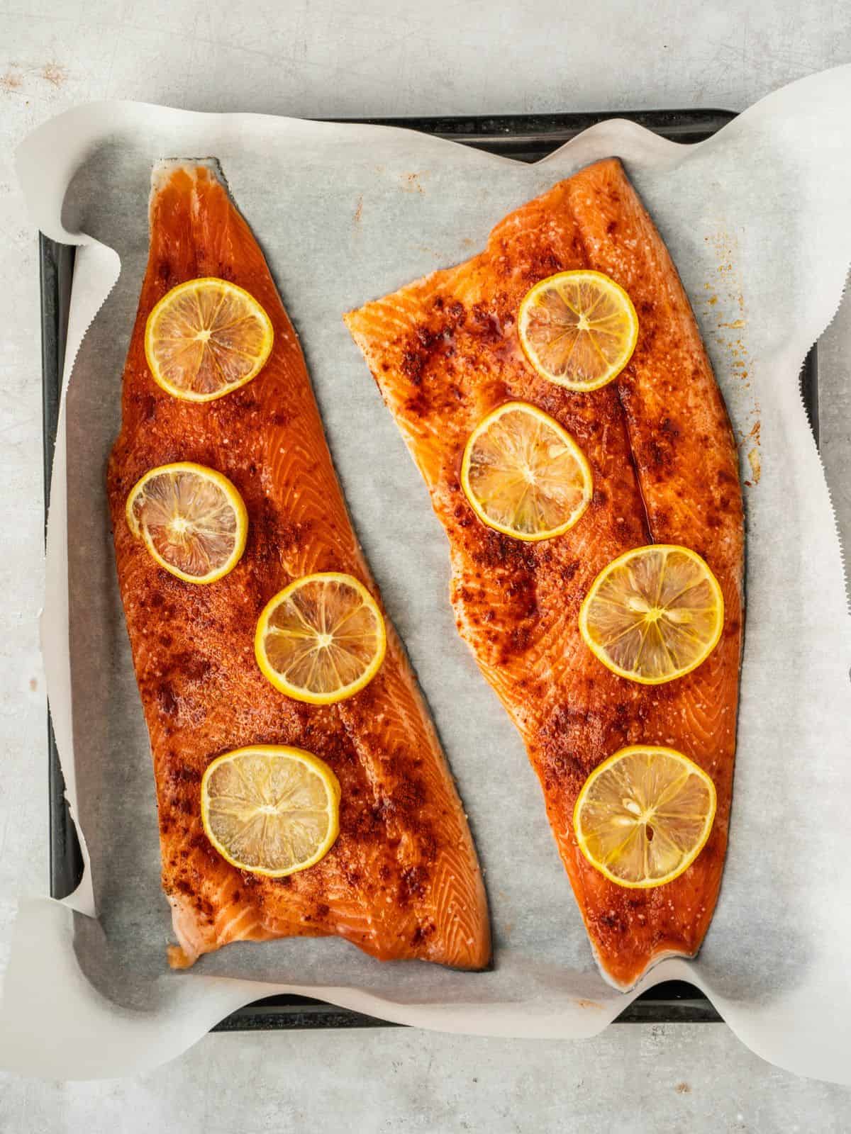 Two trout fillets on a parchment paper-lined baking sheet, seasoned with salt, pepper, paprika and lemon slices.