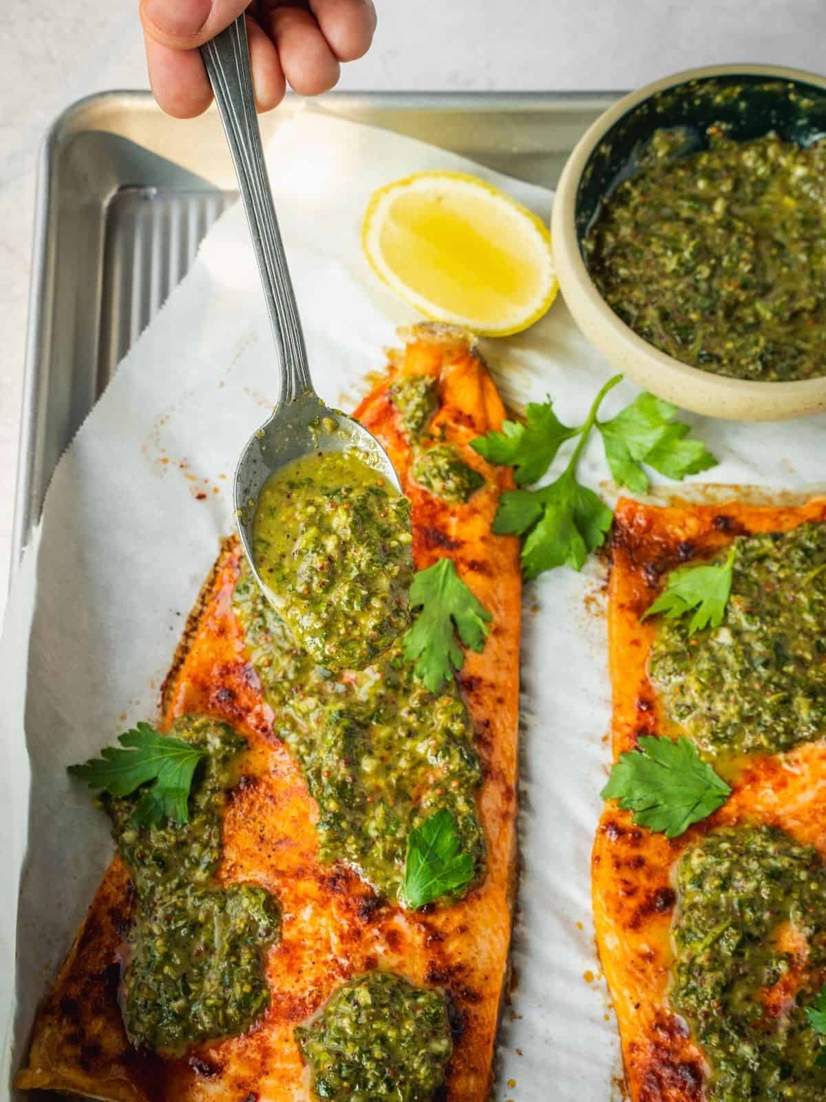 Spooning herb sauce over baked trout on a parchment paper-lined tray.