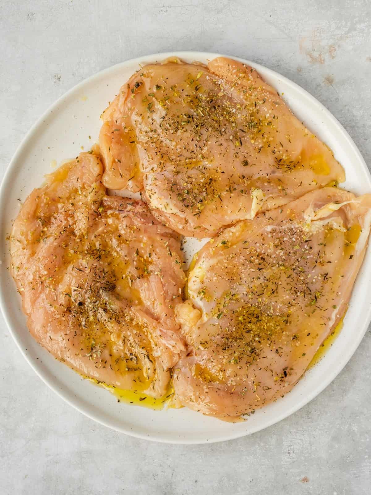 Pounded chicken breasts on a platter marinating in olive oil, oregano, salt and pepper.