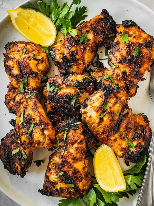 Grilled piri piri chicken thighs on a platter garnished with parsley and lemon wedges.