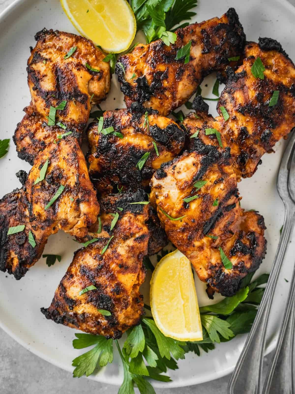 Grilled piri piri chicken thighs on a platter garnished with parsley and lemon wedges.