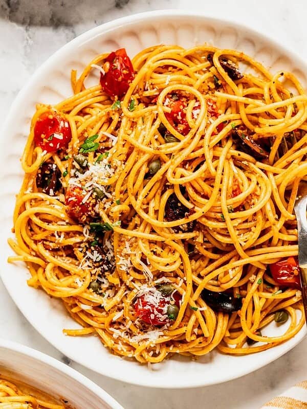 Bowl of pasta puttanesca with cherry tomatoes, garnished with cheese and chopped parsley.