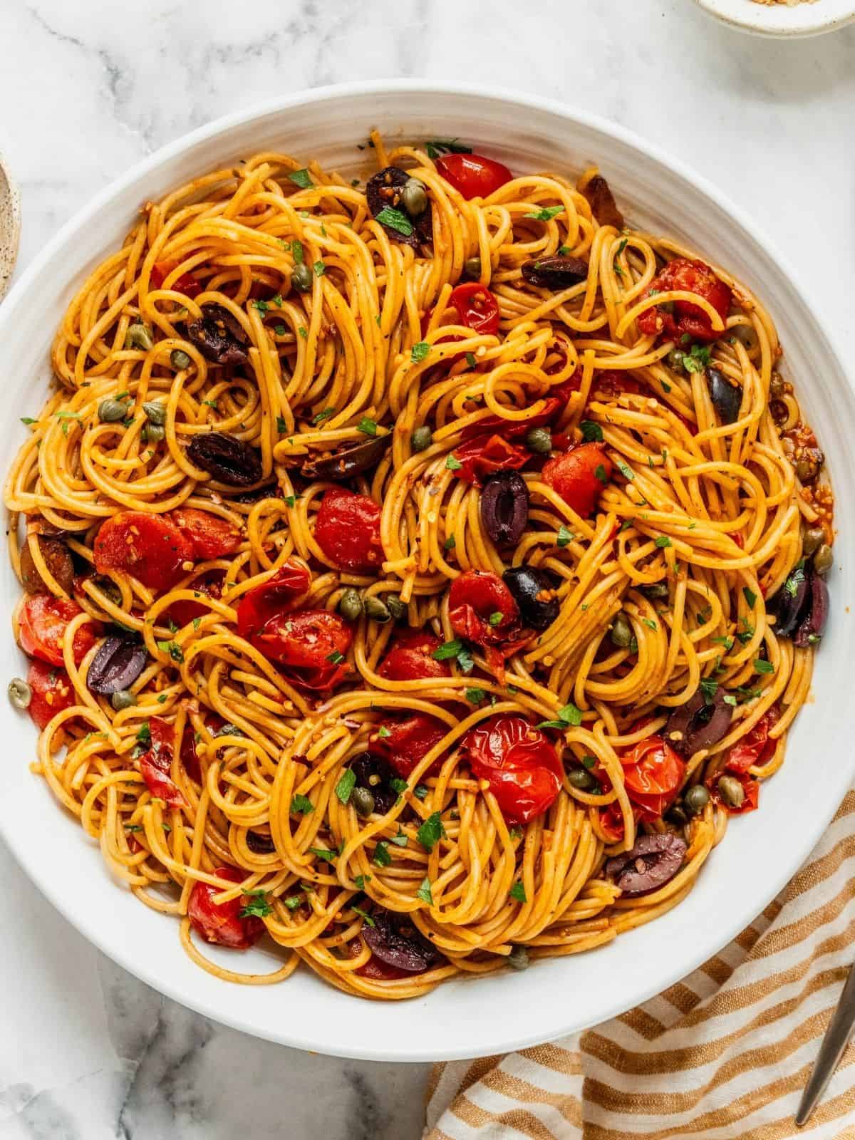 Bowl of pasta puttanesca with cherry tomatoes, capers, olives garnished with cheese and chopped parsley.
