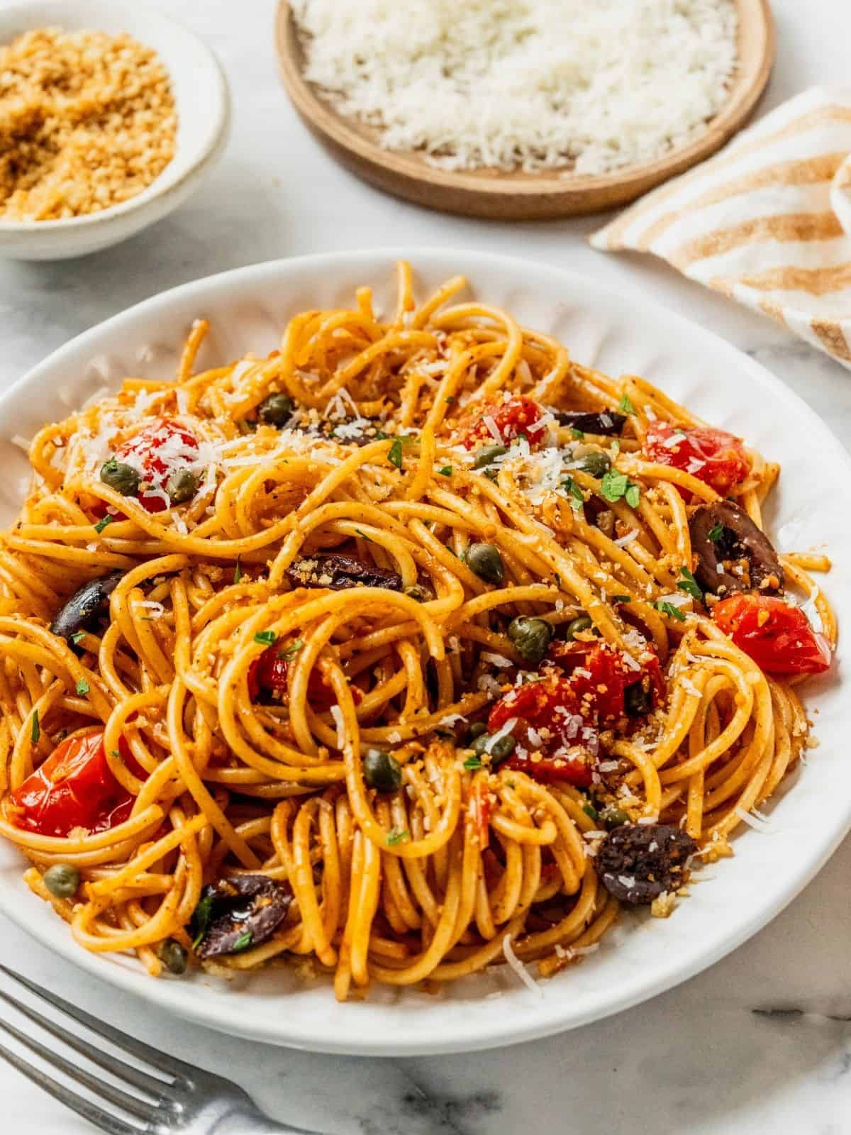 Bowl of pasta puttanesca with cherry tomatoes, capers, olives garnished with breadcrumbs, cheese and chopped parsley.