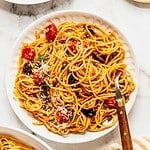 Bowl of pasta puttanesca with cherry tomatoes, garnished with cheese and chopped parsley.