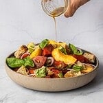 Drizzling dressing over a bowl of peach panzanella salad with chunky tomatoes, basil, croutons and shallots.