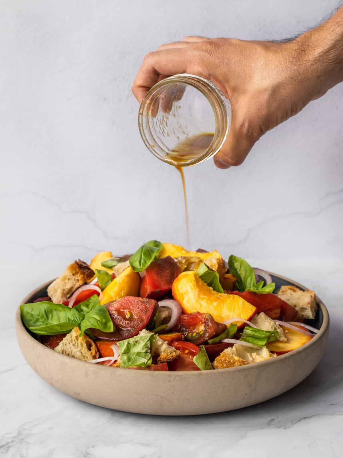 Drizzling dressing over a bowl of peach panzanella salad with chunky tomatoes, basil, croutons and shallots.