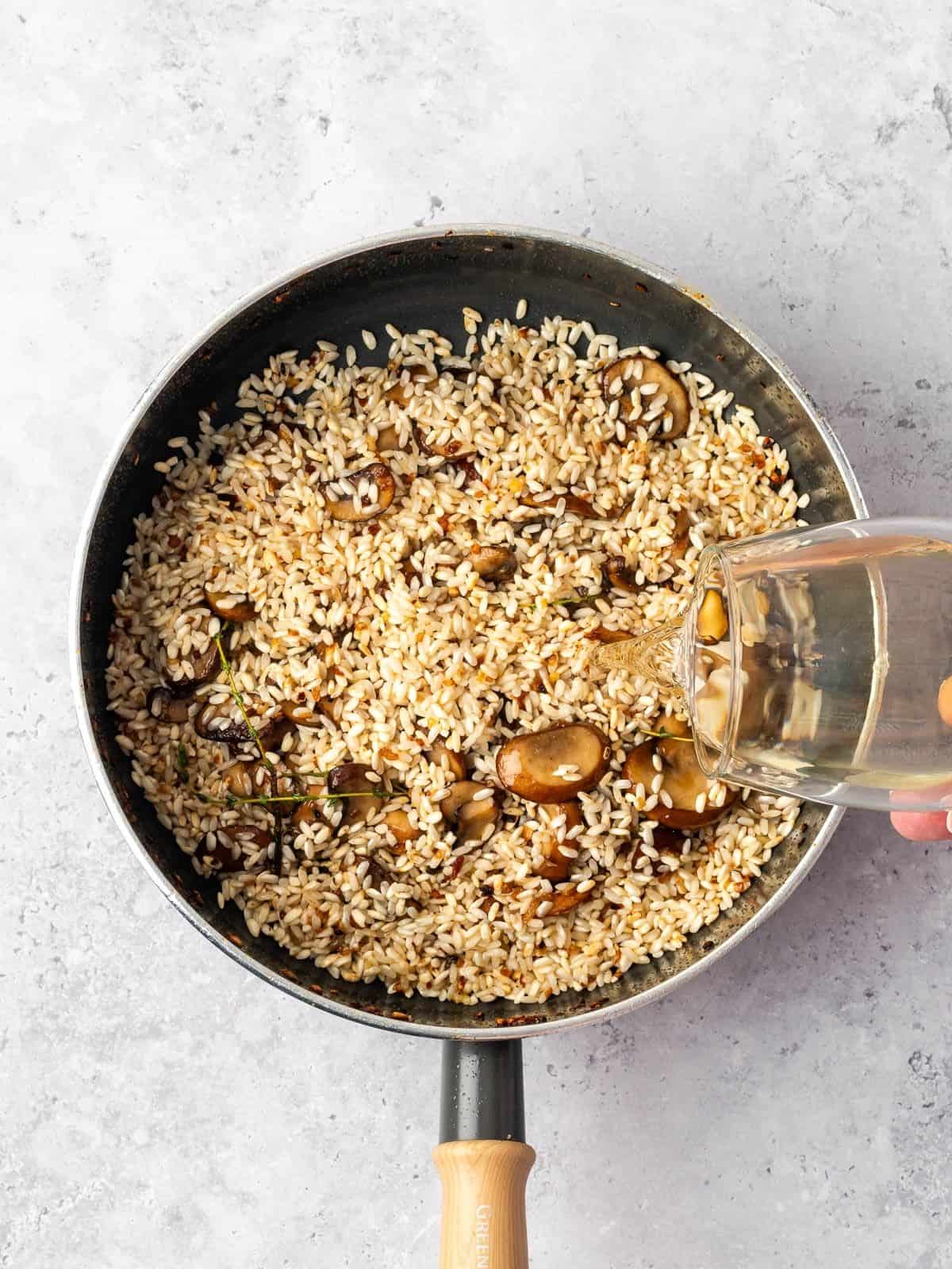 Pouring white wine into the sauté pan with the toasted rice and mushrooms.
