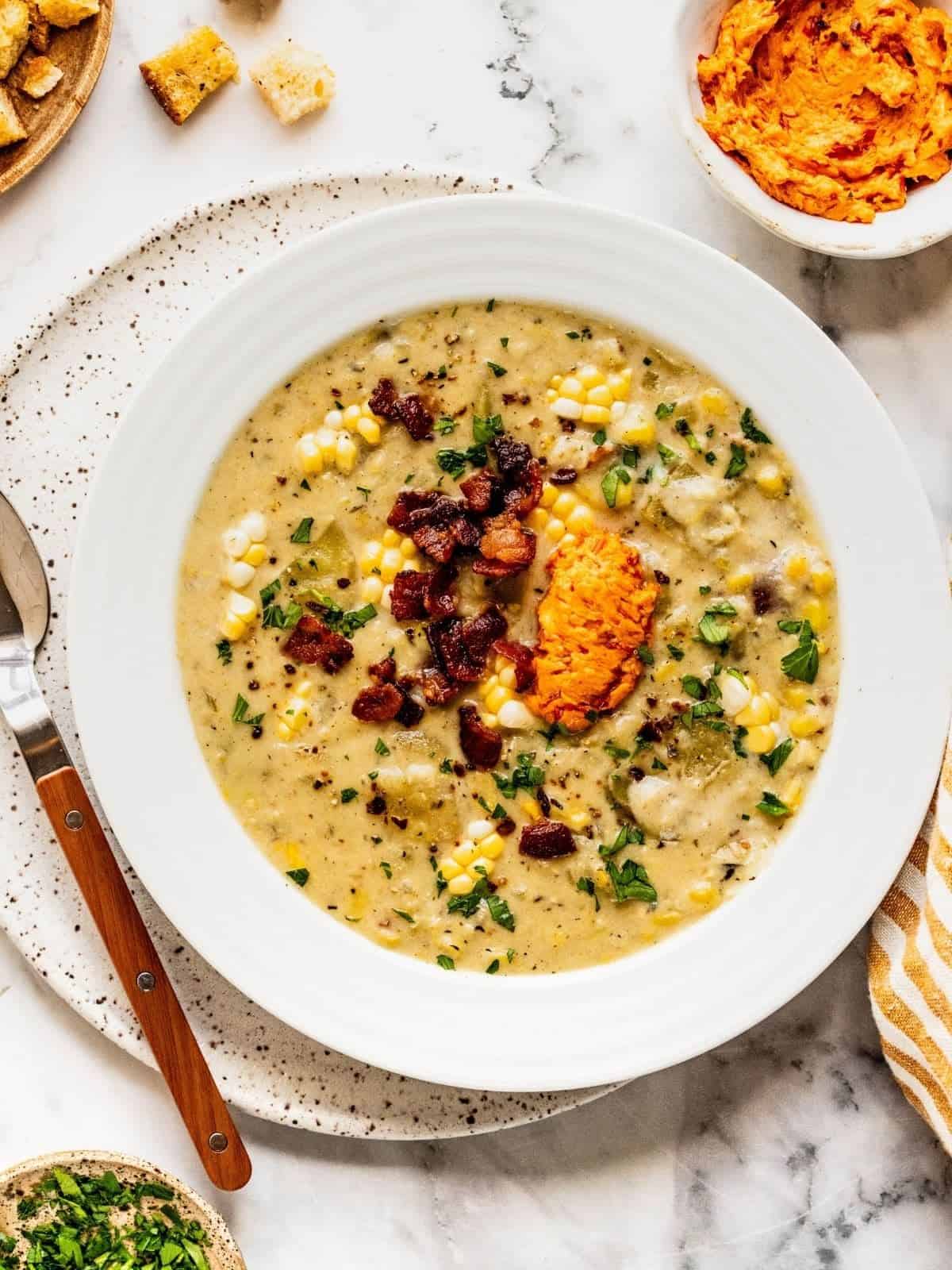 Bowl of corn chowder with harissa butter, bacon and fresh herbs.