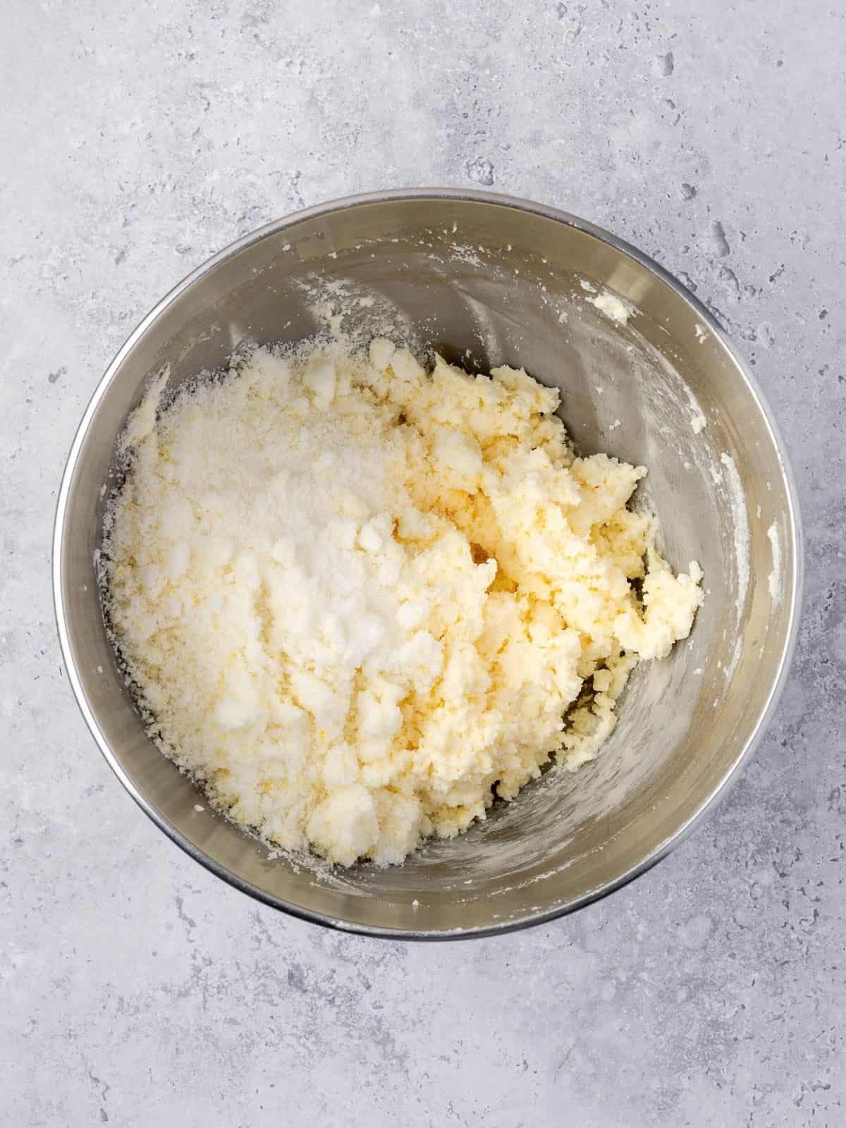 Recipe step showing the lemon sugar getting added to the whipped butter and ricotta in the bowl of a stand mixer.