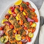 Baked Za'atar salmon fillets on a serving platter with cherry tomatoes, squash, red onion, fresh dill and lemon wedges.