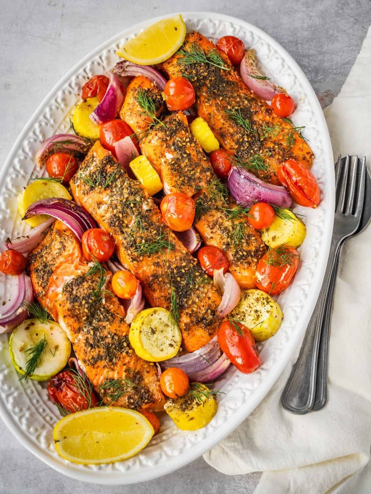 Baked Za'atar salmon fillets on a serving platter with cherry tomatoes, squash, red onion, fresh dill and lemon wedges.