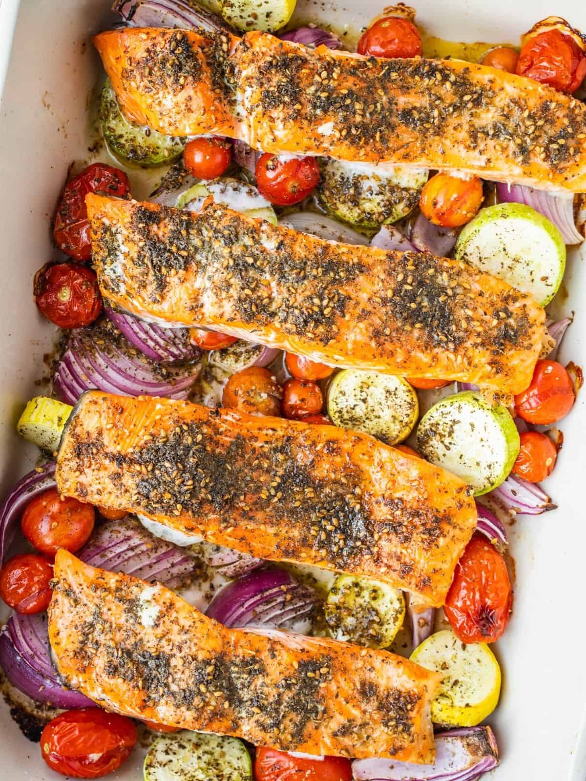 Baked Za'atar salmon fillets in a baking pan with cherry tomatoes, squash and red onion.