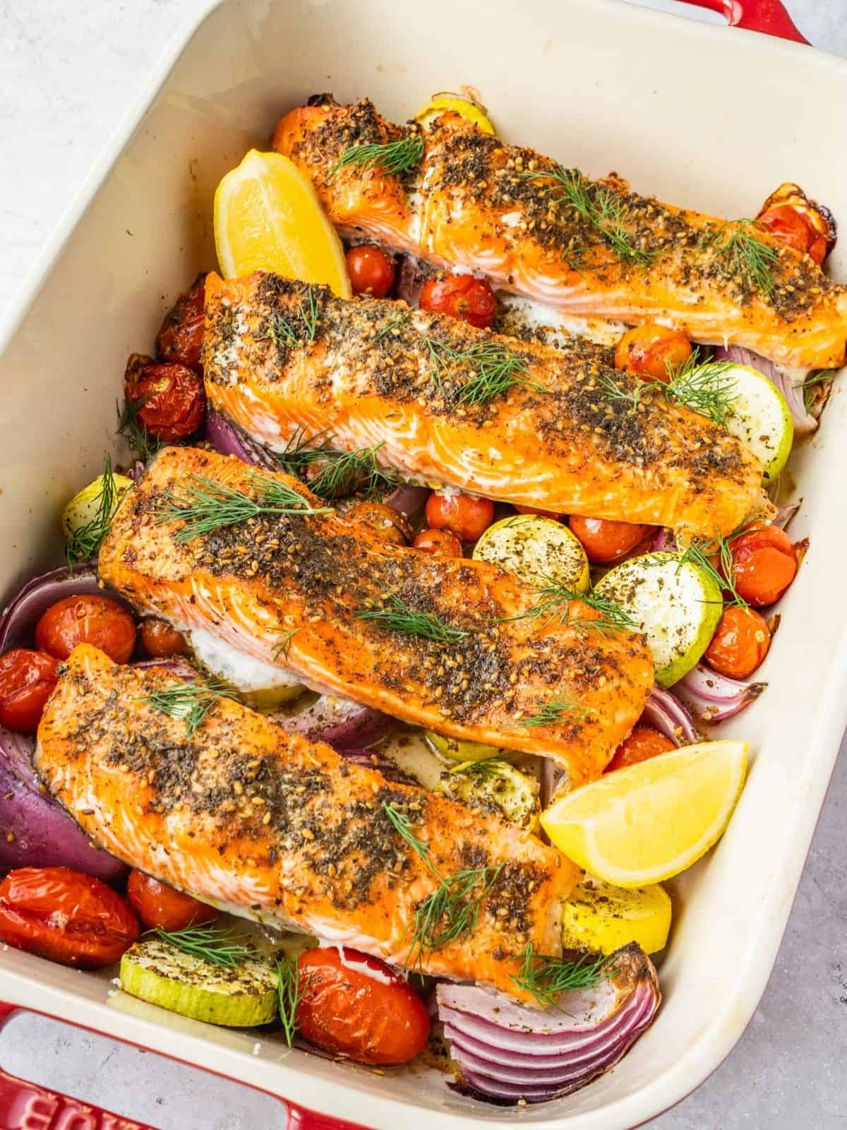 Baked Za'atar salmon fillets in a baking pan with cherry tomatoes, squash, red onion, fresh dill and lemon wedges.